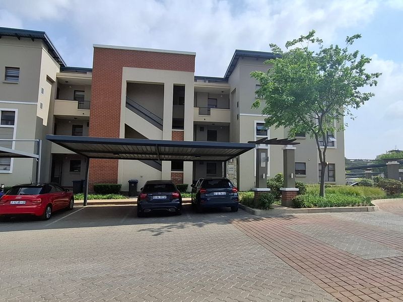 Exterior view of a modern apartment complex featuring covered parking bays, paved driveway, and well-maintained building facades with brick architectural details.