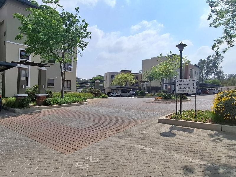 A well-maintained apartment complex entrance featuring paved driveways, covered parking bays, and established landscaped garden areas.