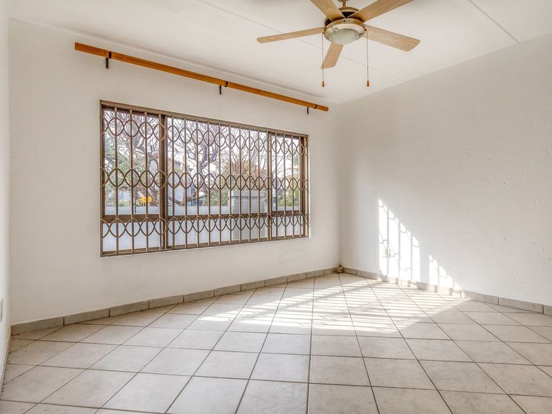 A bright, empty bedroom featuring tiled flooring, a ceiling fan, and a large window fitted with a security gate.