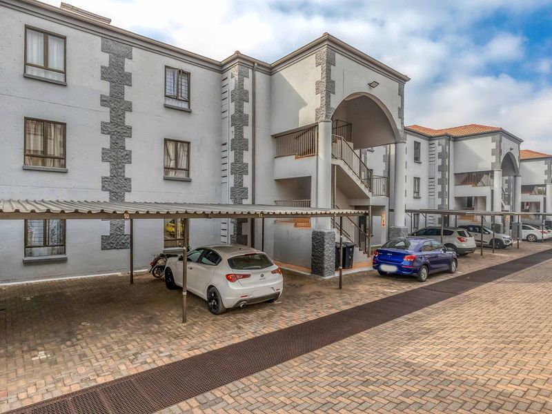 Exterior view of an apartment complex featuring covered parking bays, paved driveway, and multi-level residential units with balconies and external staircases.