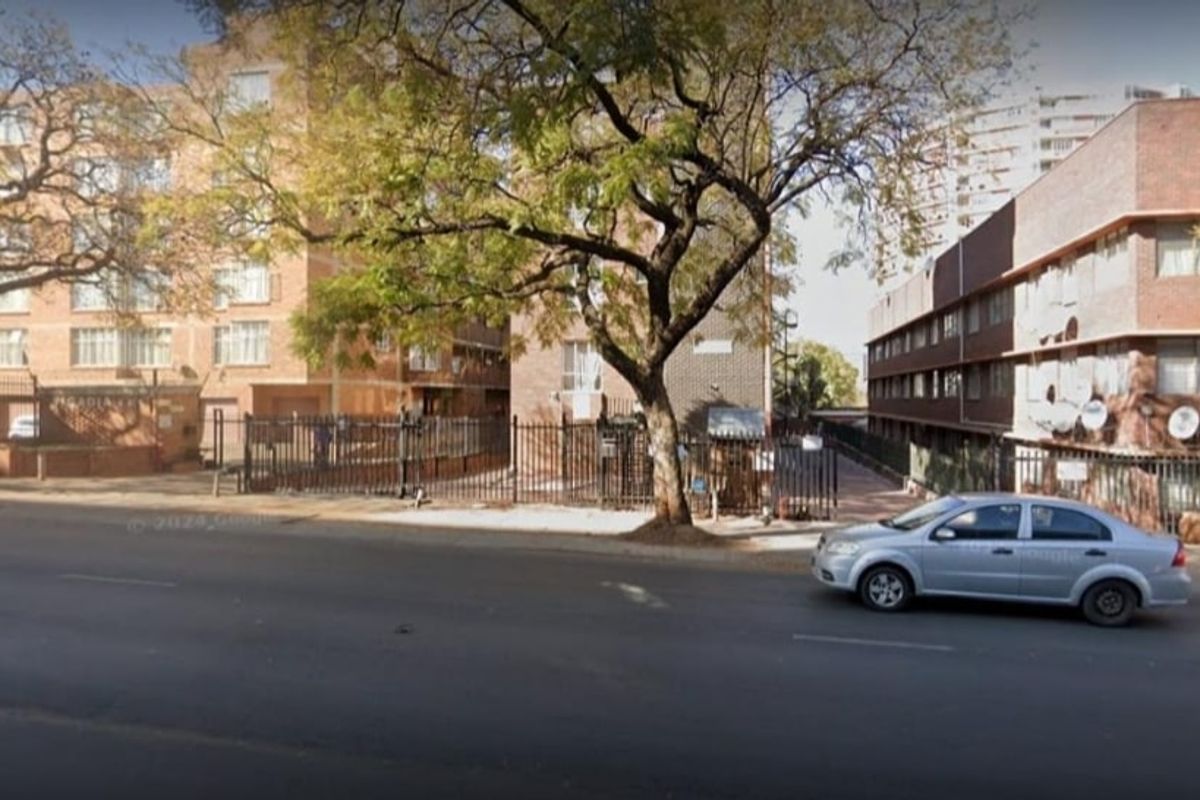 Street-level view of the apartment complex exterior, featuring brick building facades, security fencing, and mature trees along the sidewalk.