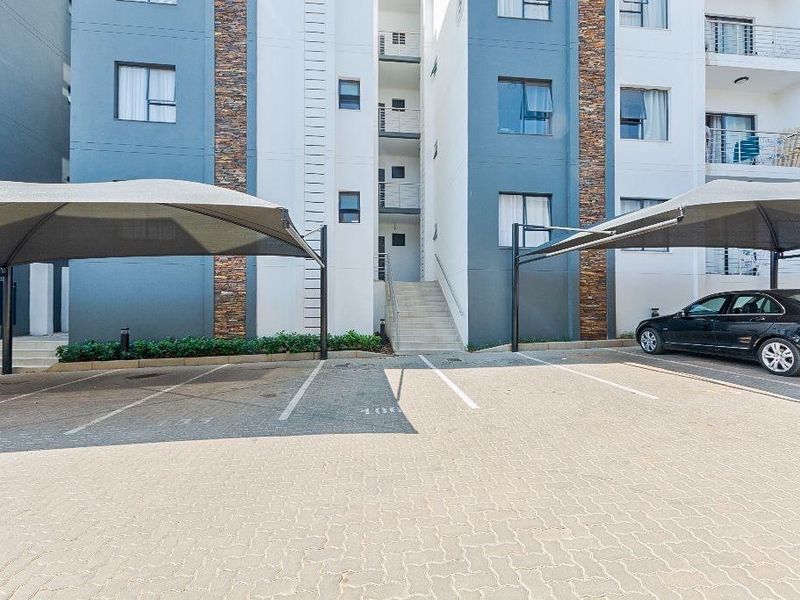 Front view of the apartment complex featuring paved parking bays with shade cloth carports and modern stone-clad building facade.