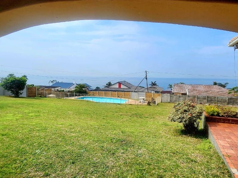 Covered patio overlooking a spacious lawn, swimming pool, and distant ocean views, providing a functional outdoor entertainment space.