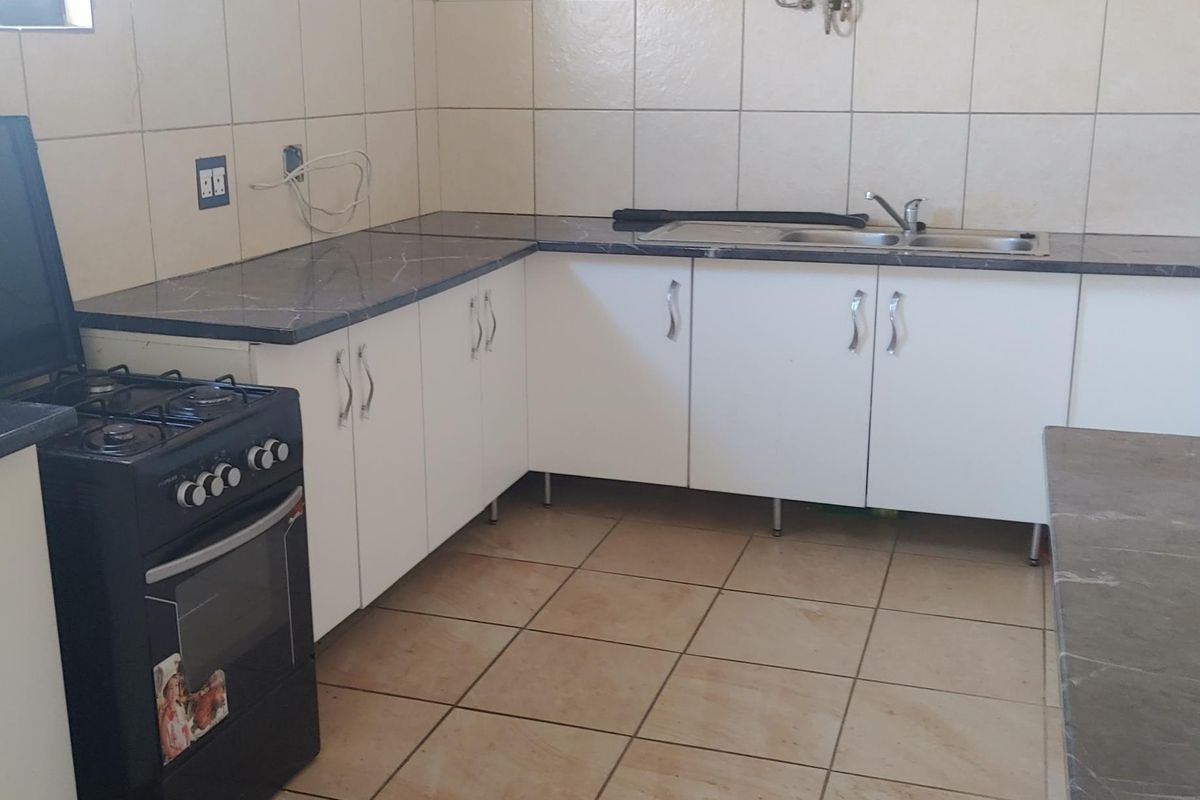 A functional kitchen featuring white cabinetry, dark laminate countertops, tiled backsplash, and a gas geyser, requiring some cosmetic updates to the walls and finishes.
