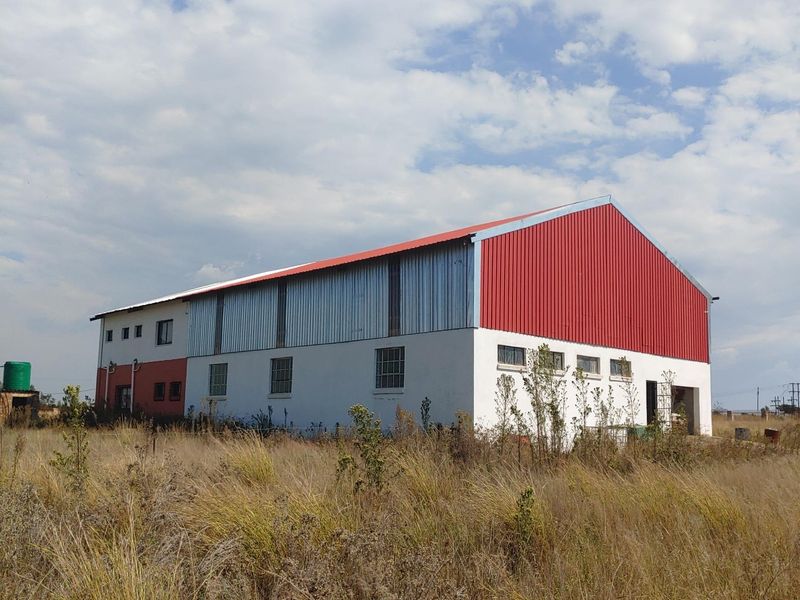A large industrial-style building featuring a red corrugated metal roof, metal cladding, and plastered walls, situated on a large plot with overgrown grass.