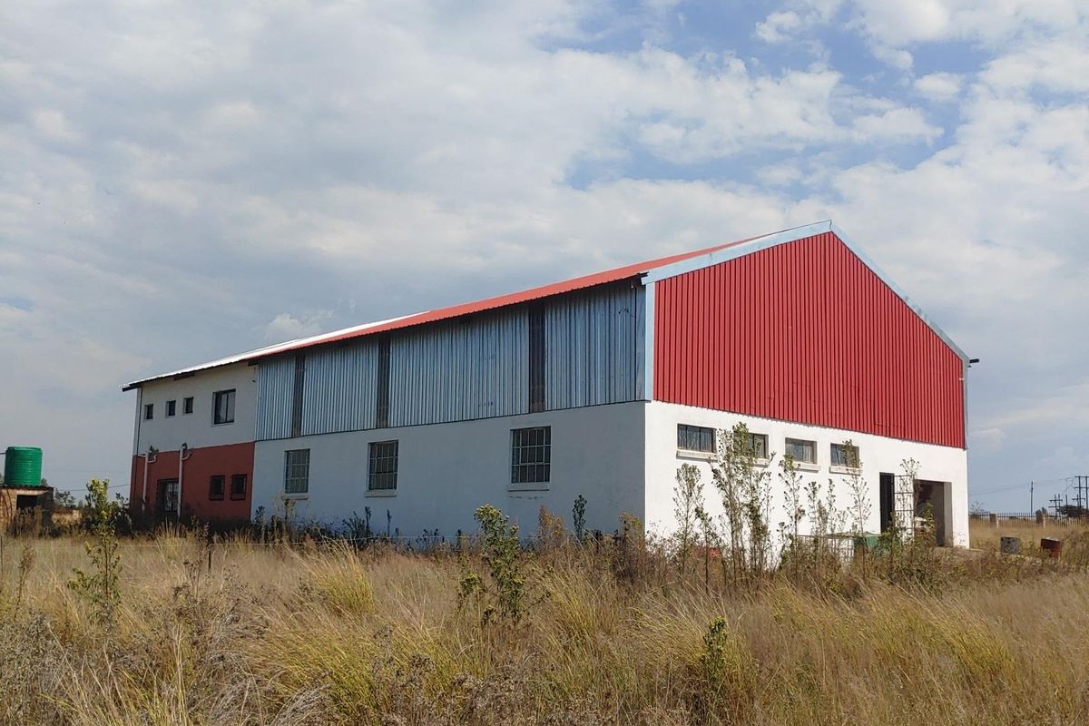 A large industrial-style building featuring a red corrugated metal roof, metal cladding, and plastered walls, situated on a large plot with overgrown grass.