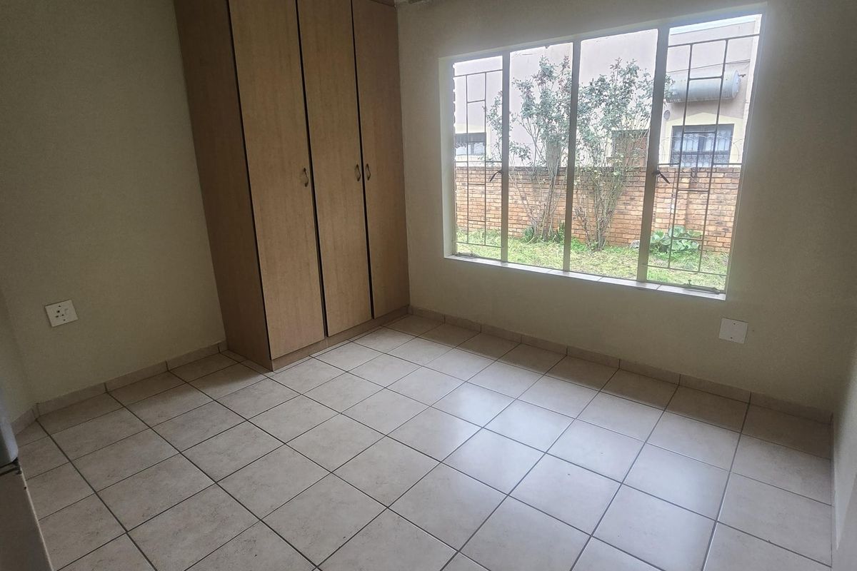 A clean, empty bedroom featuring neutral tiled flooring, a built-in wooden wardrobe, and a large window with security bars overlooking the garden.