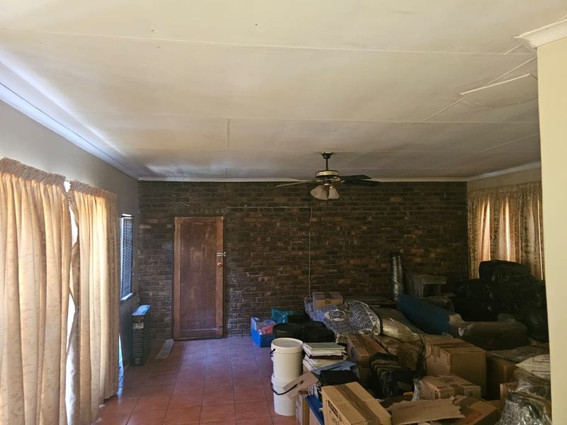 A living room featuring an exposed brick feature wall, tiled flooring, and a ceiling fan, currently cluttered with storage items.