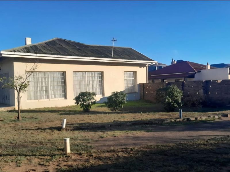 Front view of a single-story house featuring a corrugated iron roof, large windows, and a spacious yard with a brick boundary wall.