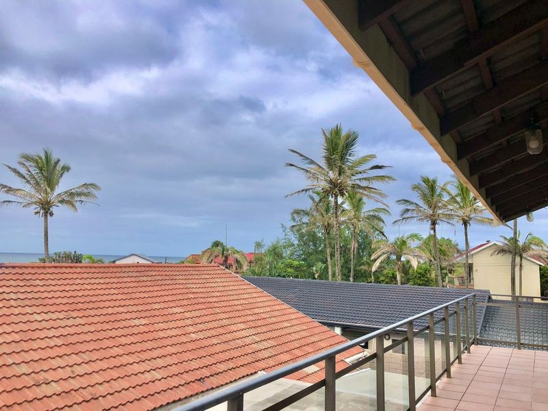 A covered balcony featuring tiled flooring and a modern glass balustrade, offering elevated views of the surrounding neighborhood and distant ocean.