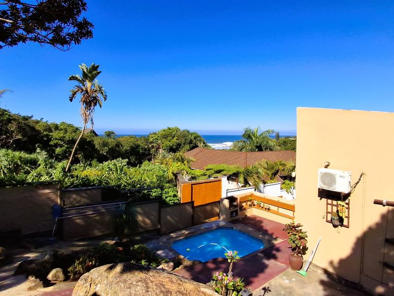 Outdoor pool area featuring a built-in braai, paved patio, and scenic coastal views, set within a landscaped garden environment.