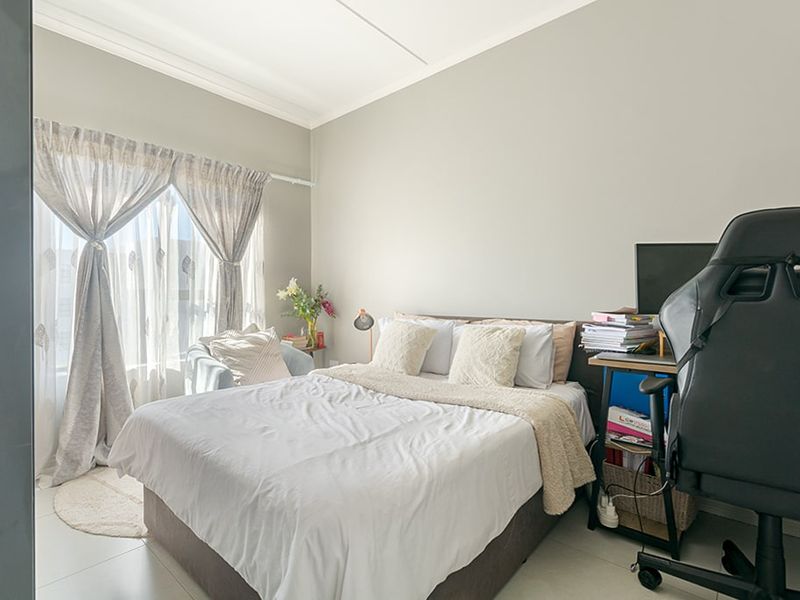 A well-lit bedroom featuring neutral wall tones, tiled flooring, and large windows with curtain rails, providing a comfortable and functional living space.