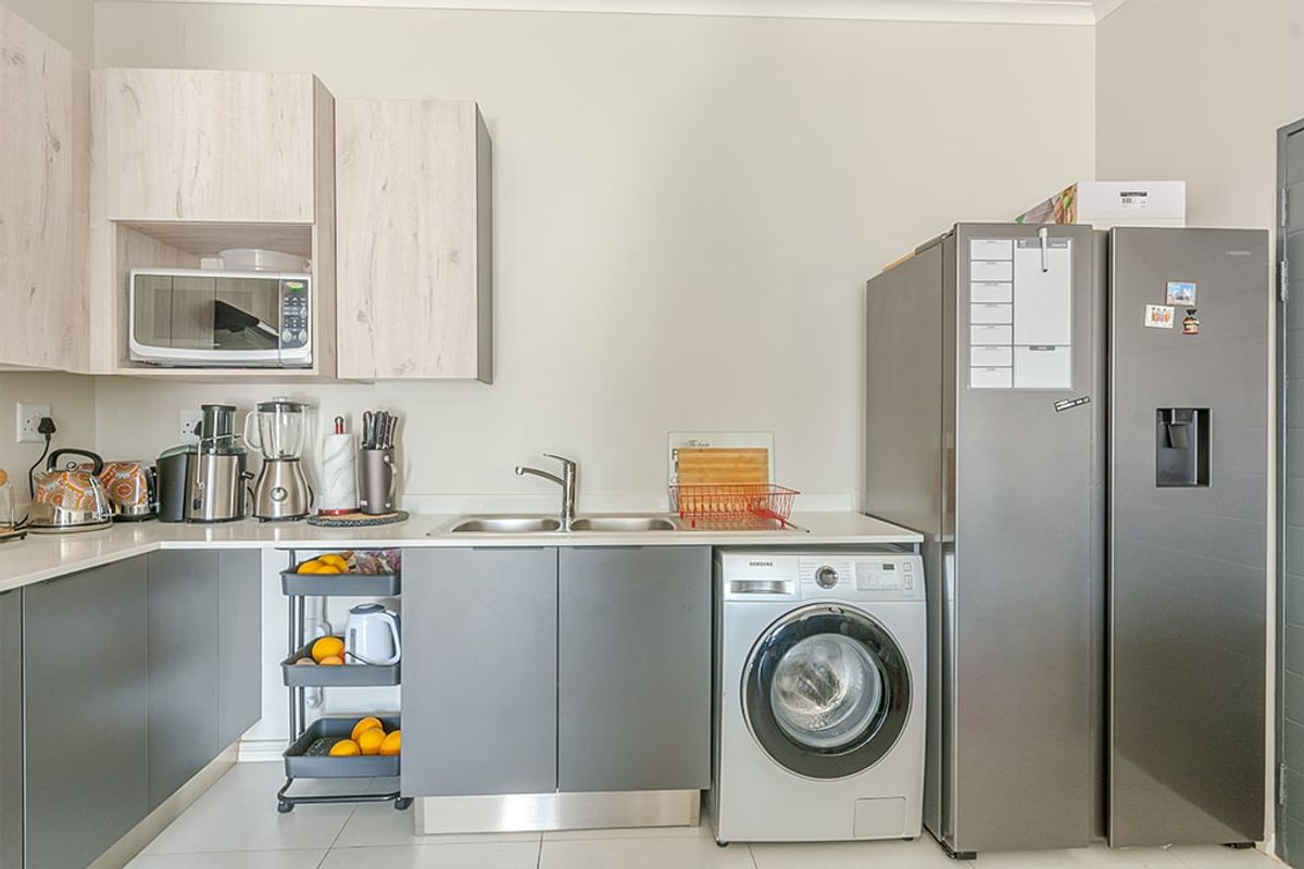 A modern kitchen featuring light wood-grain upper cabinets, dark grey lower cabinetry, a stainless steel sink, and integrated space for laundry appliances.