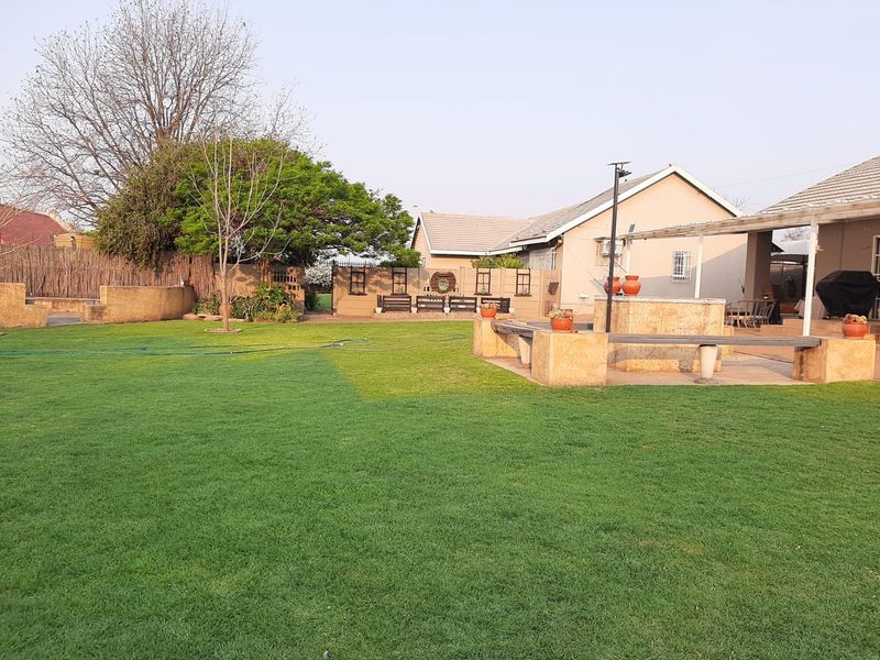 Spacious rear garden featuring a well-maintained lawn, a paved patio area, and a built-in stone braai with integrated bench seating.