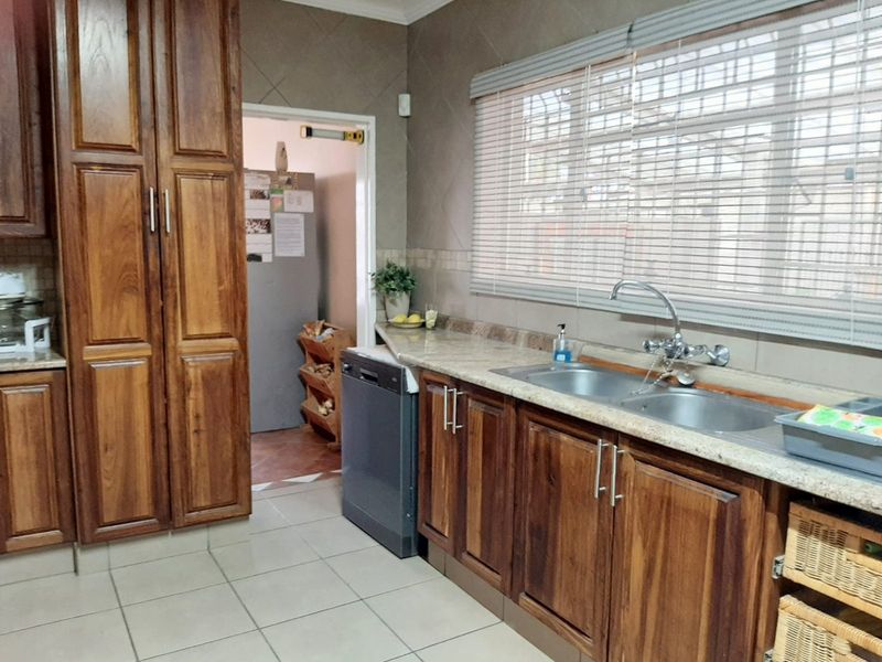 A functional kitchen featuring warm-toned wooden cabinetry, granite countertops, a double stainless steel sink, and tiled flooring with a view into a scullery area.