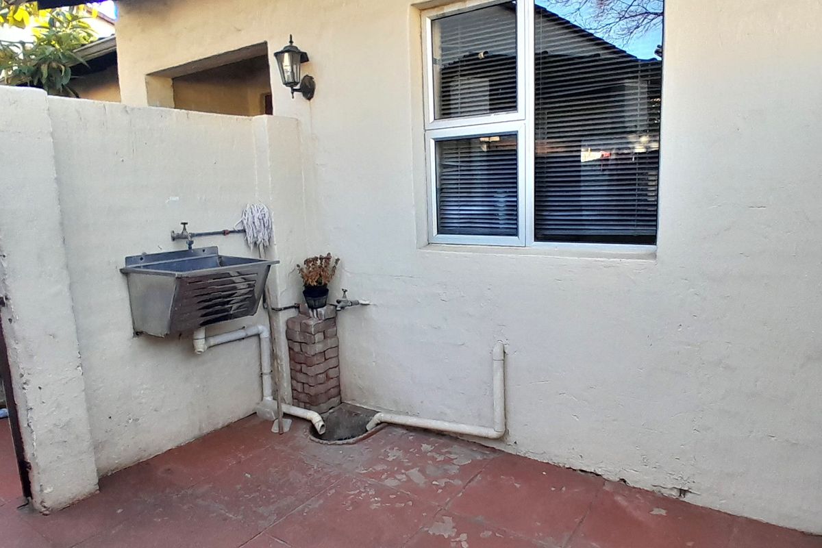 Rear exterior utility area featuring a stainless steel sink, tiled flooring, and exposed plumbing fixtures against a plastered wall.