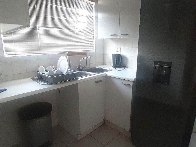 A compact kitchen featuring white built-in cabinetry, a stainless steel sink, tiled backsplash, and tiled flooring with natural light from the window.