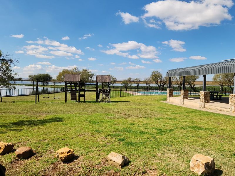 Spacious landscaped garden featuring a swimming pool, covered patio with stone-clad pillars, and playground equipment, overlooking a scenic waterfront.