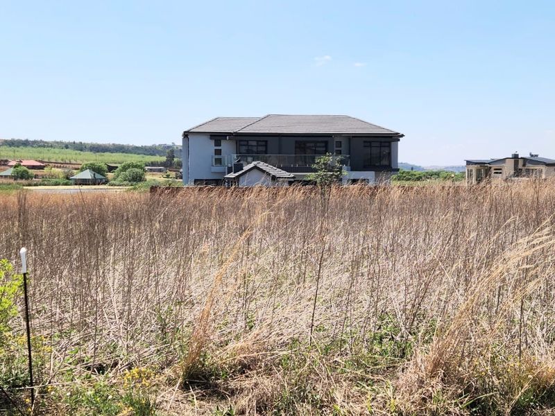 A view of a vacant residential plot with tall dry grass, featuring a neighboring modern house in the background.