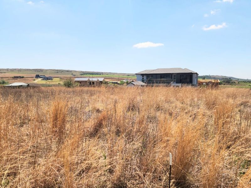 A view of a vacant residential plot featuring dry grass, with established and under-construction homes visible in the surrounding neighborhood.