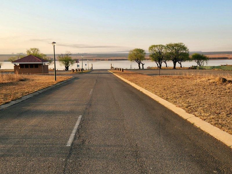 A paved access road leading towards a waterfront area, featuring concrete curbing, street lighting, and a small brick utility structure.