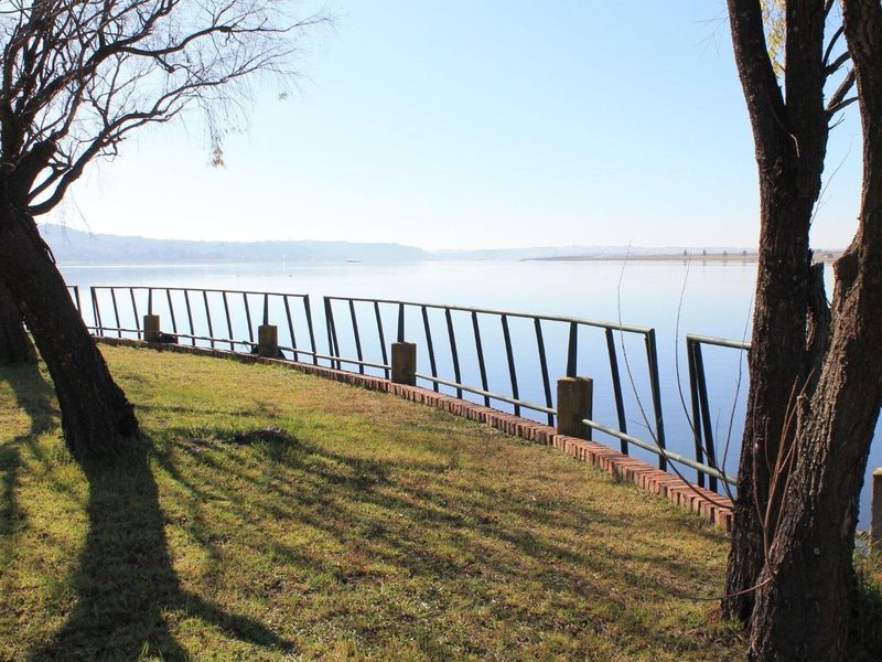 A scenic waterfront view featuring a grassy bank, brick retaining wall, and metal perimeter fencing overlooking a calm lake.