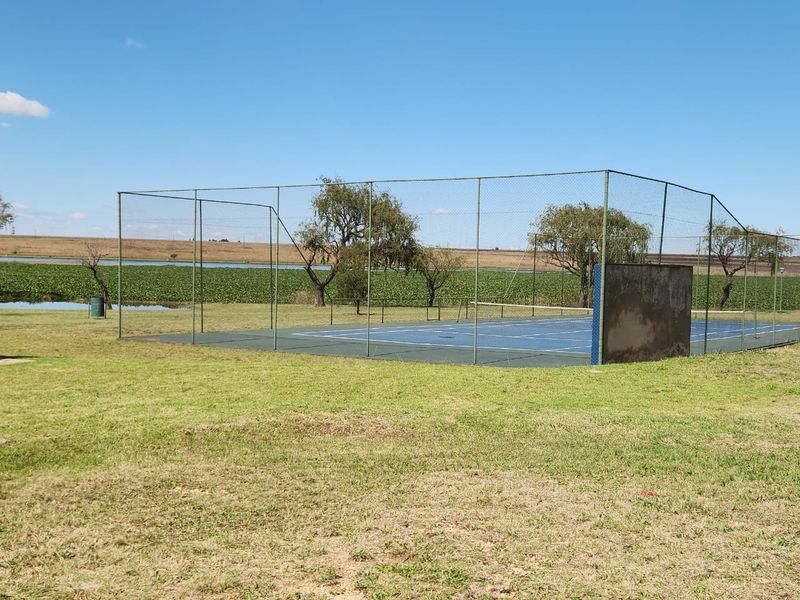 Outdoor tennis court facility featuring a blue hard court surface, perimeter chain-link fencing, and a practice wall, set within a grassy landscape.