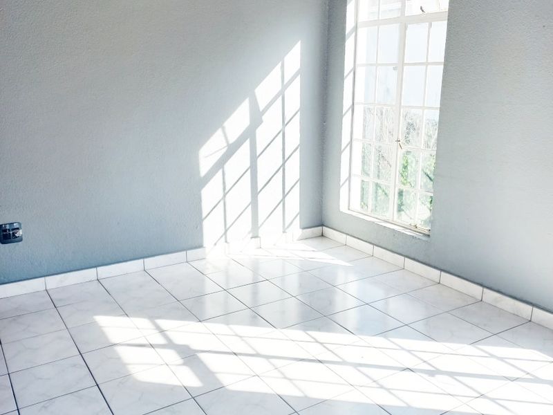 A bright, empty living area featuring white tiled flooring, exposed timber ceiling beams, and a large grid-pattern window allowing ample natural light.