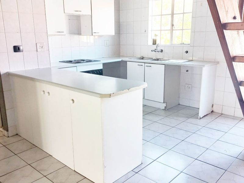 A dated kitchen featuring white cabinetry, tiled surfaces, and an integrated hob and oven, adjacent to a wooden staircase in an open-plan apartment layout.