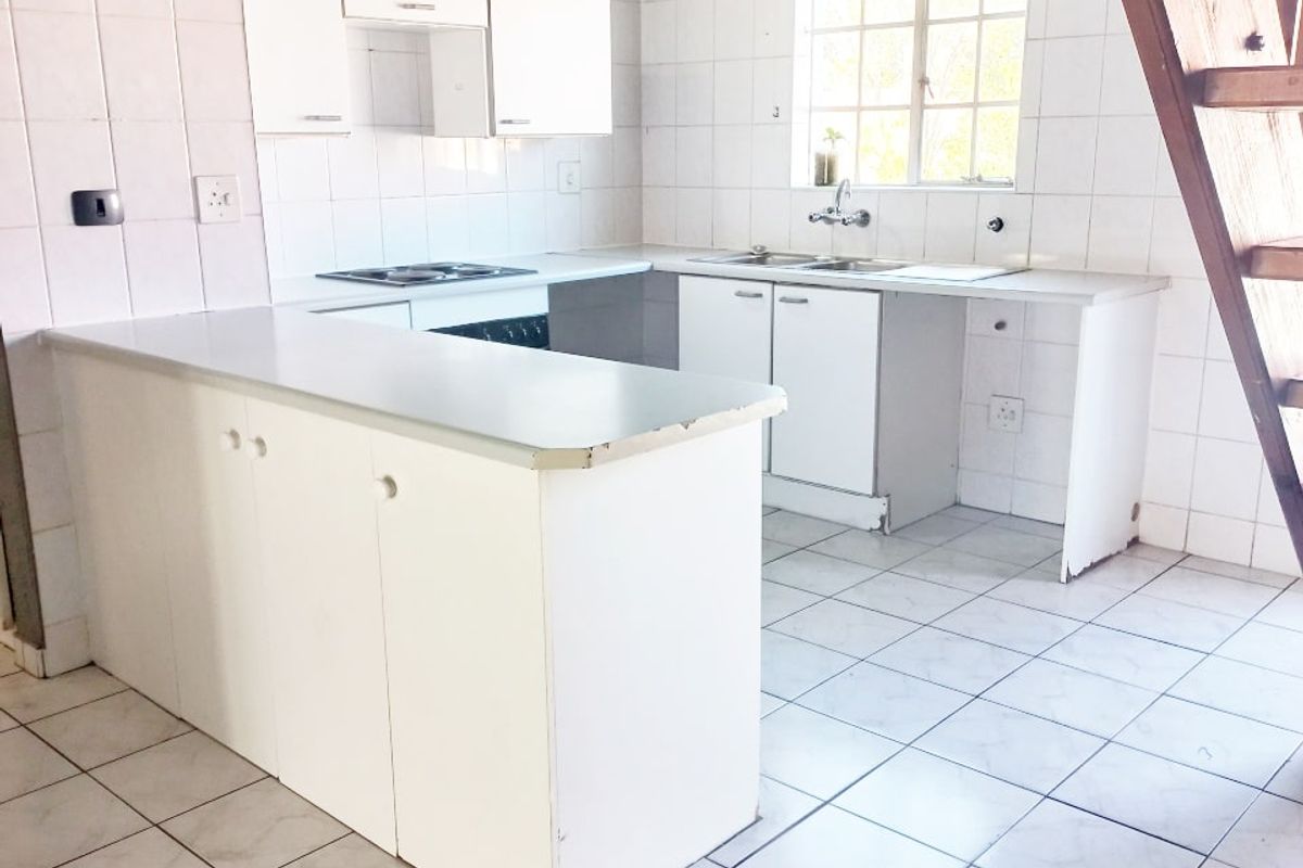 A dated kitchen featuring white cabinetry, tiled surfaces, and an integrated hob and oven, adjacent to a wooden staircase in an open-plan apartment layout.