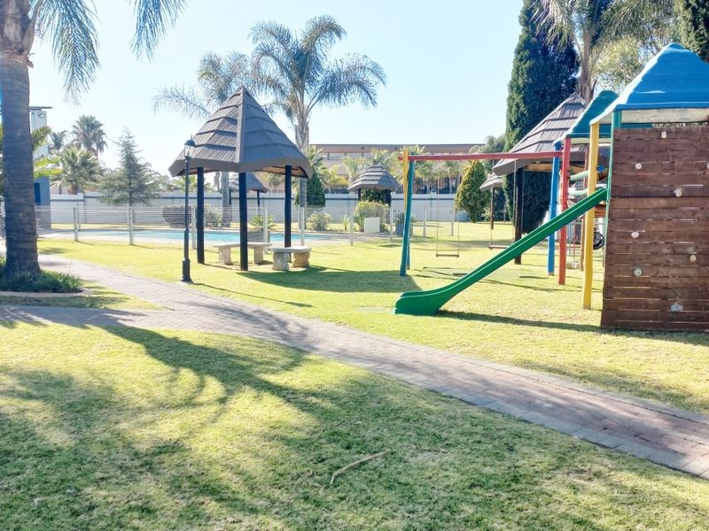A well-maintained communal garden area featuring a paved walkway, a thatched gazebo, and children's playground equipment set within a landscaped lawn.