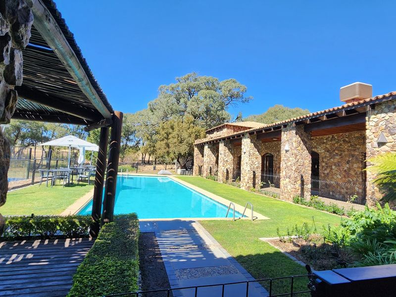 A well-maintained outdoor pool area featuring a stone-clad building, timber-covered patio, and landscaped garden surroundings.