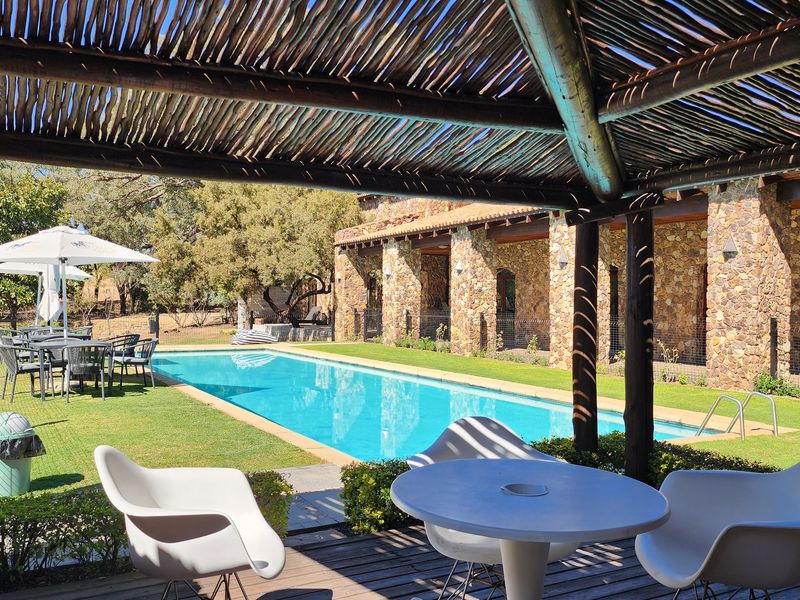 A well-maintained outdoor pool area featuring a stone-clad building, a thatched pergola, and a clear swimming pool set within a landscaped garden.