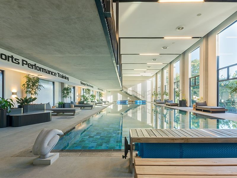 A modern indoor swimming pool area featuring floor-to-ceiling windows, tiled pool surround, and recessed lighting, offering a bright and professional training environment.