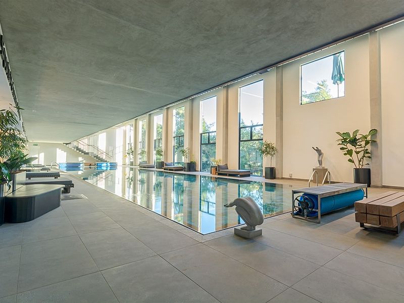 A spacious indoor swimming pool area featuring large format floor tiles, a concrete ceiling, and expansive full-height windows providing natural light.