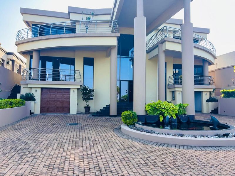 A grand multi-level modern residence featuring a paved driveway, a decorative water feature, and prominent structural columns leading to the main entrance.