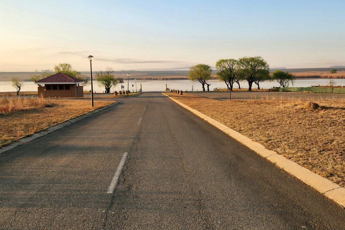 A paved access road leading towards a waterfront area, featuring concrete curbing, street lighting, and a small brick utility structure.
