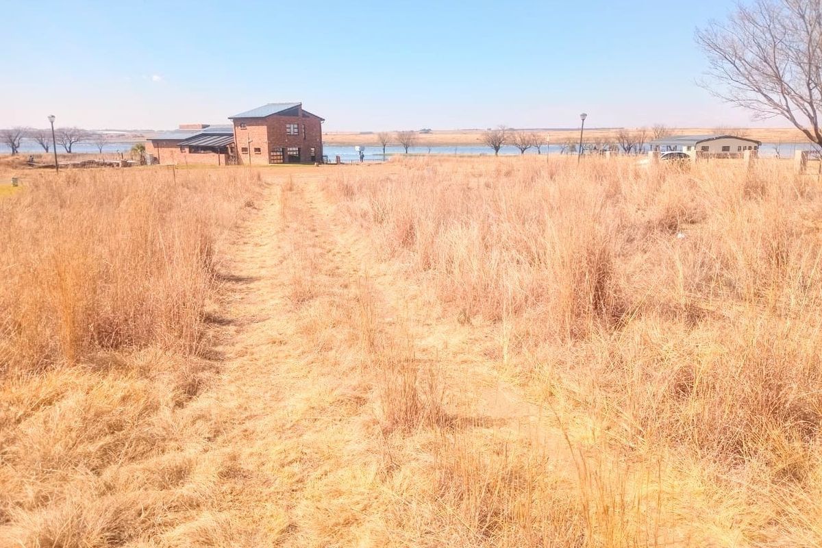 A view of a vacant residential plot featuring dry grass, an unpaved access path, and distant views of existing estate buildings and water.