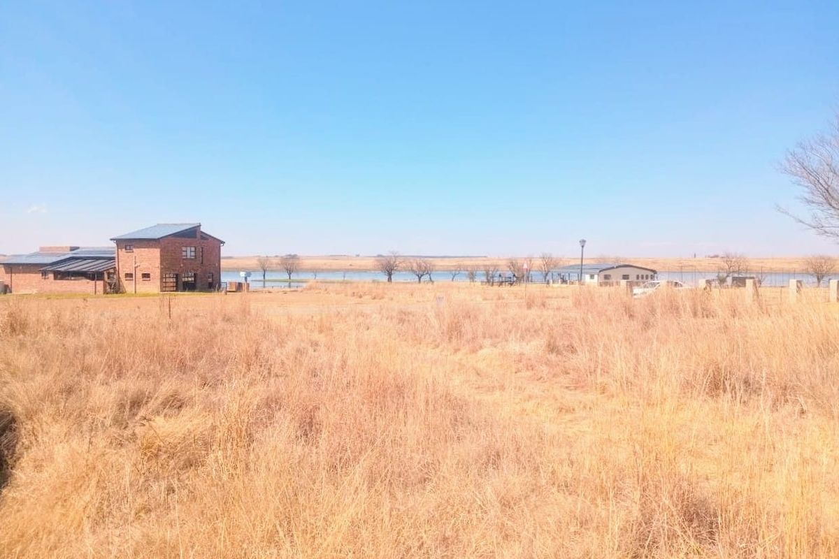 A wide-angle view of a vacant residential plot featuring dry grass, distant buildings, and a waterfront landscape.