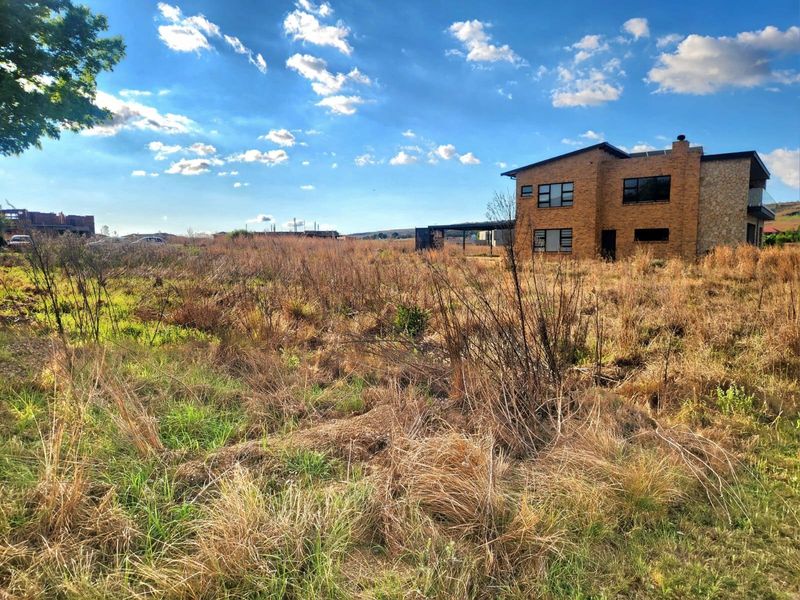A view of a vacant residential plot featuring overgrown grass with an adjacent multi-story brick house visible in the background.