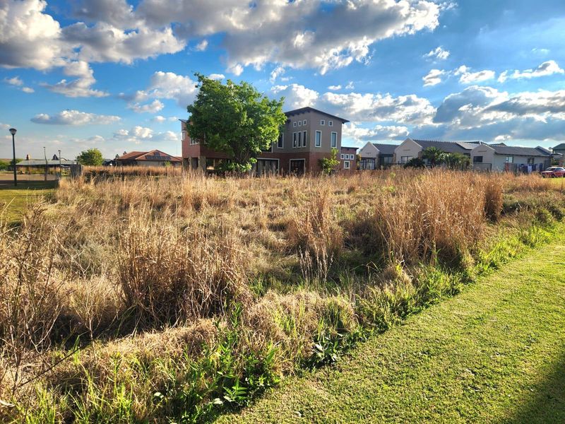 A view of a vacant residential plot featuring overgrown grass, situated in a developed neighborhood with surrounding residential properties.