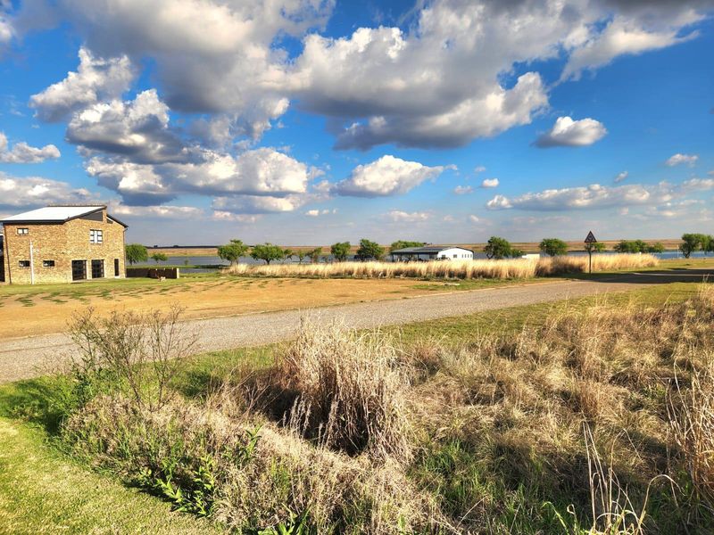 A wide-angle view of a vacant residential plot featuring a gravel driveway, open grassy landscape, and a nearby brick building under a clear sky.