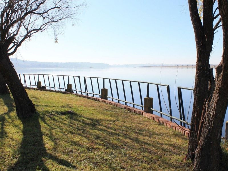 A scenic waterfront view featuring a grassy bank, a brick retaining wall, and a damaged metal safety railing overlooking the calm water.