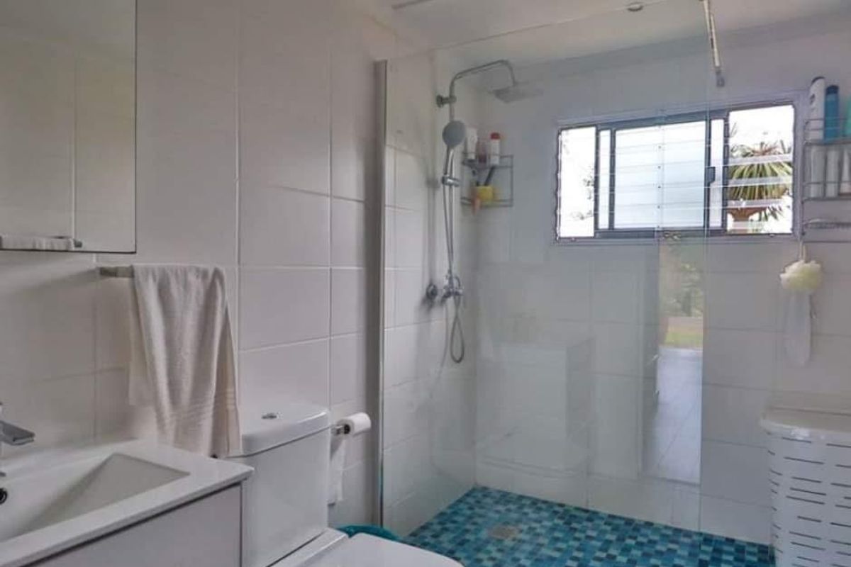A clean, functional bathroom featuring a vanity cabinet, a glass-enclosed shower with mosaic floor tiling, and a louver window for natural ventilation.