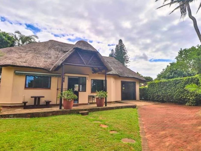Front view of a townhouse featuring a traditional thatch roof, a covered patio area, a brick-paved driveway, and a well-maintained garden.