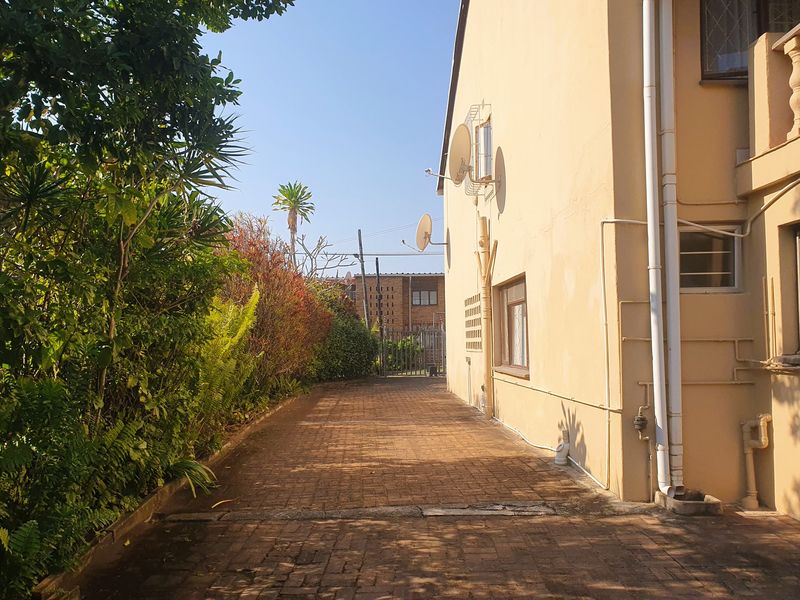 A side exterior view of the property featuring a paved walkway, established garden borders, and external building infrastructure including downpipes and satellite dishes.