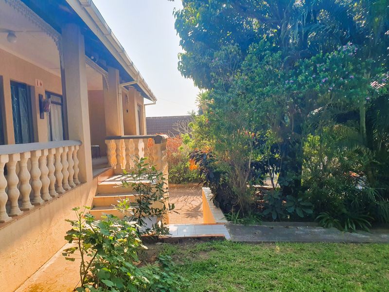 A rear exterior view featuring a covered veranda with a concrete balustrade, leading down to a paved patio area surrounded by established garden greenery.