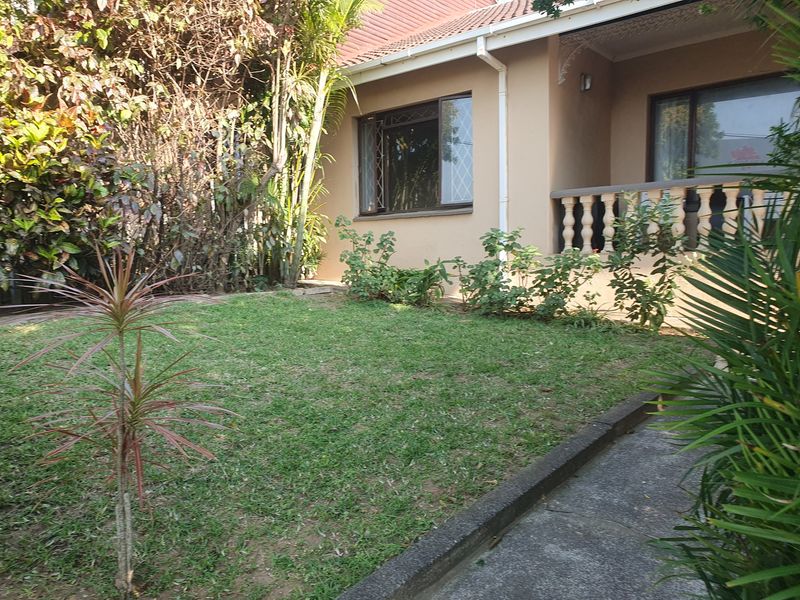 A rear garden view featuring a lawn area, concrete pathway, and a covered patio with a balustrade attached to the main house structure.