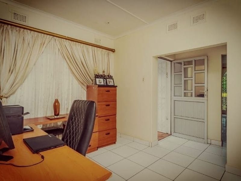 A functional home office space featuring tiled flooring, neutral walls, and direct access to an adjacent room through a panelled door.