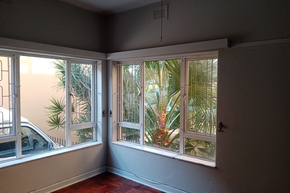 An empty living room featuring classic parquet flooring, large corner windows with security bars, and natural light, offering a blank canvas for interior design.